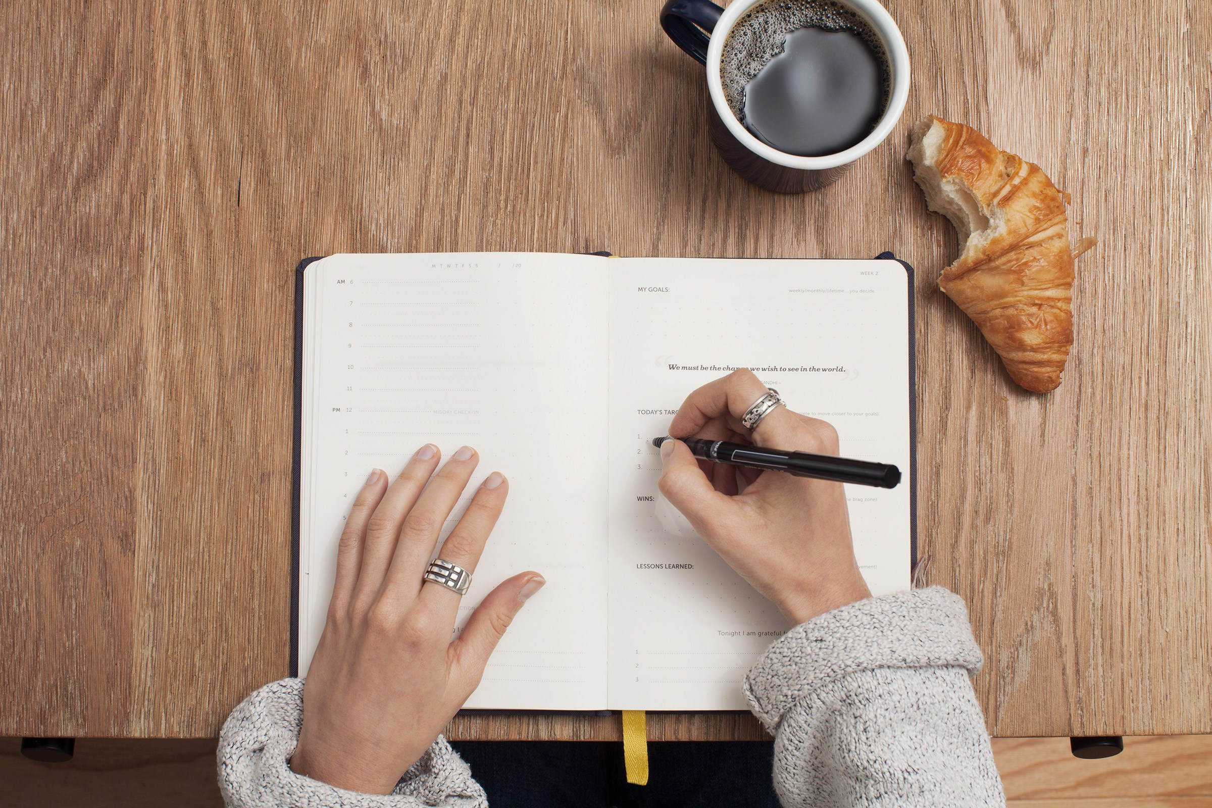 Person writing in a notebook with a cup of coffee nearby on a wooden table.